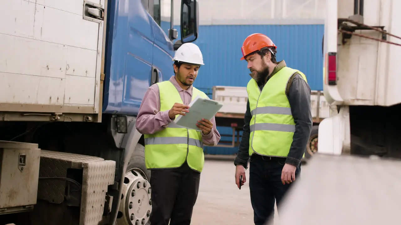 Boss And Worker Wearing Vests And Safety Helmets Organizing A Truck Fleet In A Logistics Park While They Consulting A Document 5