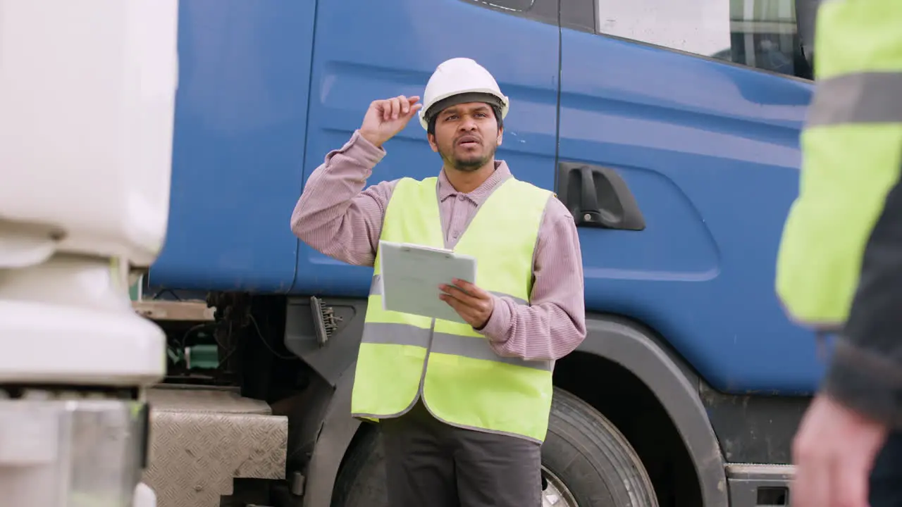 Distant View Of Worker Wearing Vest And Safety Helmet Organizing A Truck Fleet In A Logistics Park While Reading A Document