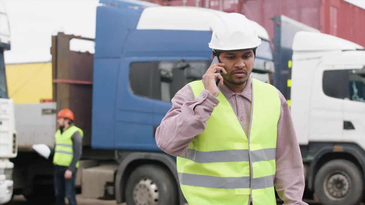 Worker Wearing Vest And Safety Helmet Organizing A Truck Fleet In A Logistics Park While Talking On The Phone 1