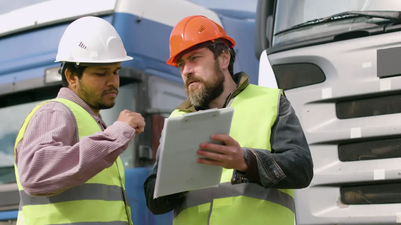 Worker Wearing Vest And Safety Helmet Organizing A Truck Fleet In A Logistics Park While Consulting A Document And Holding A Smartphone 1