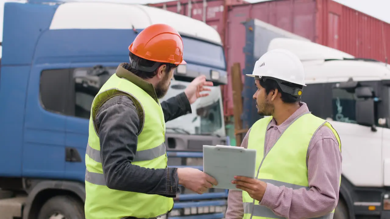 Boss And Worker Wearing Vests And Safety Helmets Organizing A Truck Fleet In A Logistics Park While They Consulting A Document