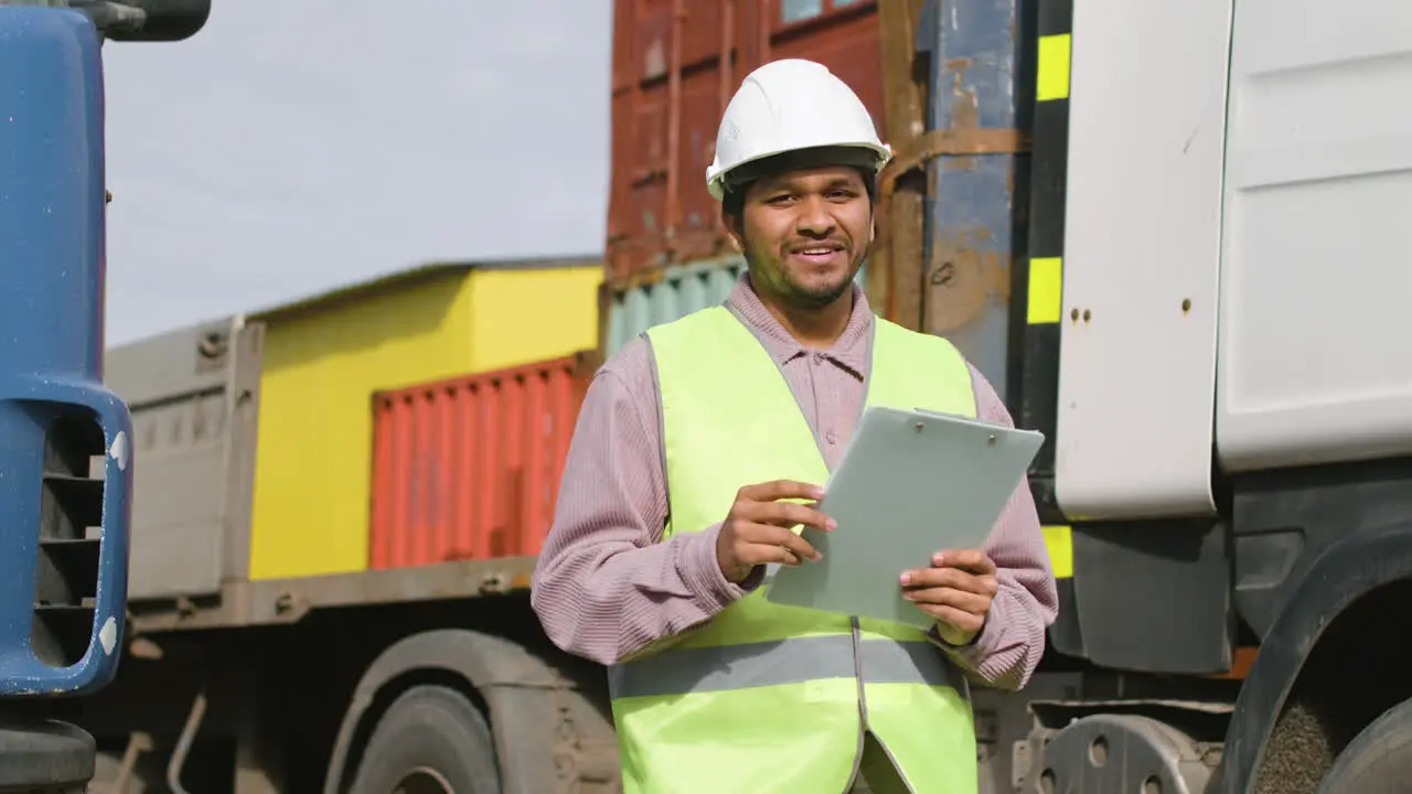 Worker Wearing Vest And Safety Helmet Looking And Smiling At Camera In A Logistics Park While Holding Documents 1