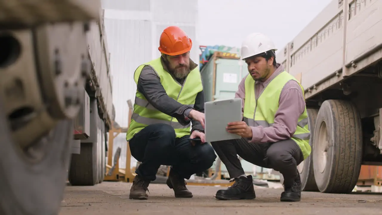 Boss And Worker Wearing Vests And Safety Helmets Organizing A Truck Fleet Squatting In A Logistics Park While They Reading A Document