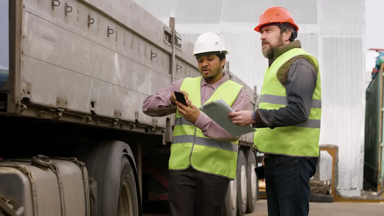 Boss And Worker Wearing Vests And Safety Helmets Organizing A Truck Fleet In A Logistics Park While They Reading On A Smartphone And Document