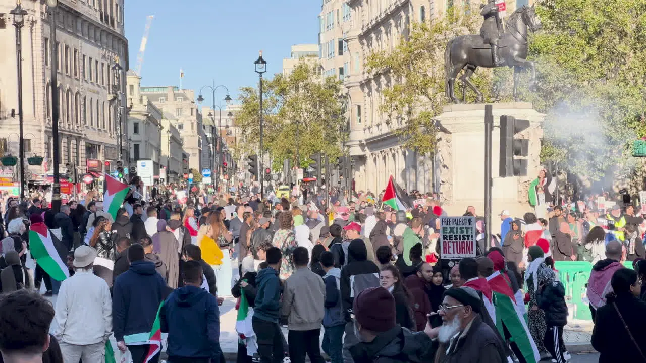 Large crowd marches down city street with flags and banners under clear skies London UK