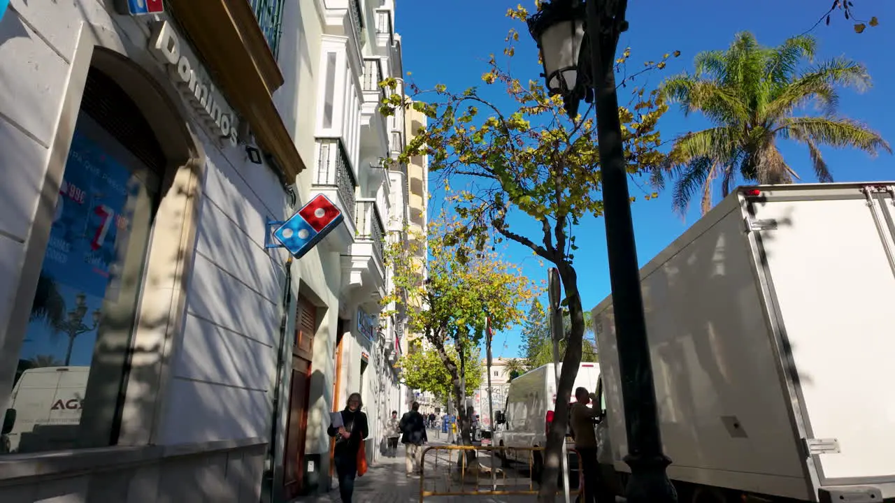 A quaint Cádiz street featuring traditional architecture with a bright advertisement banner against a backdrop of blue sky and treetops