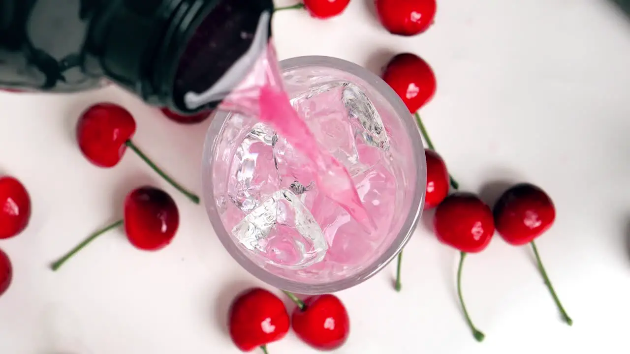 Close up birdseye shot of a shaker pouring a tasty cherry whey to clear ice cubes while cherries lie on the side