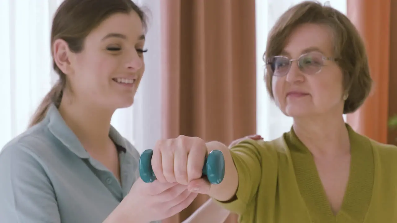 Elder Woman Holding A Dumbbell Raising An Arm Up Assisted By A Female Doctor 3