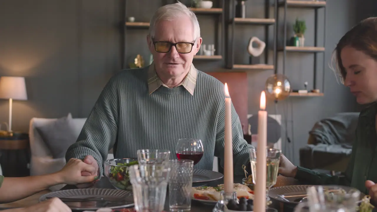 Old Man And His Two Adult Daughters Holding Hands And Praying Before Meal While Sitting At Dinner Table At Home