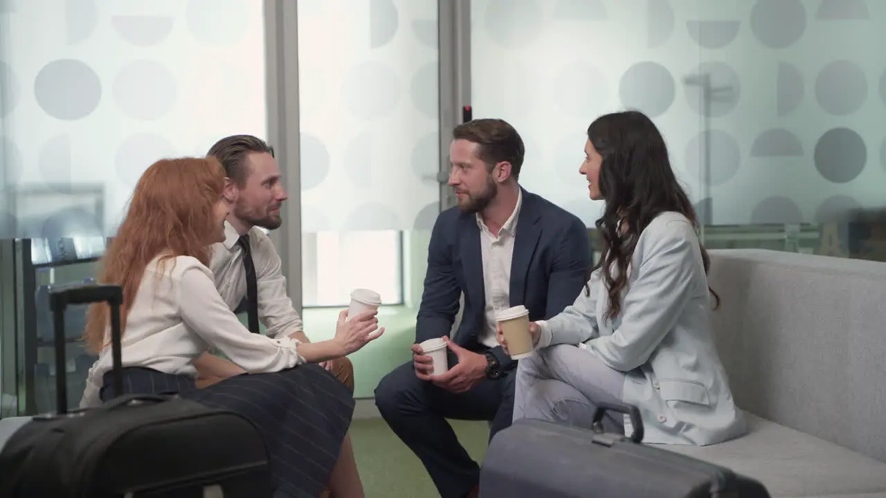 A Working Group Of Two Businesswomen And Two Businessmen Chatting Relaxedly With Some Coffees In The Lobby Of An Airport Before A Business Trip