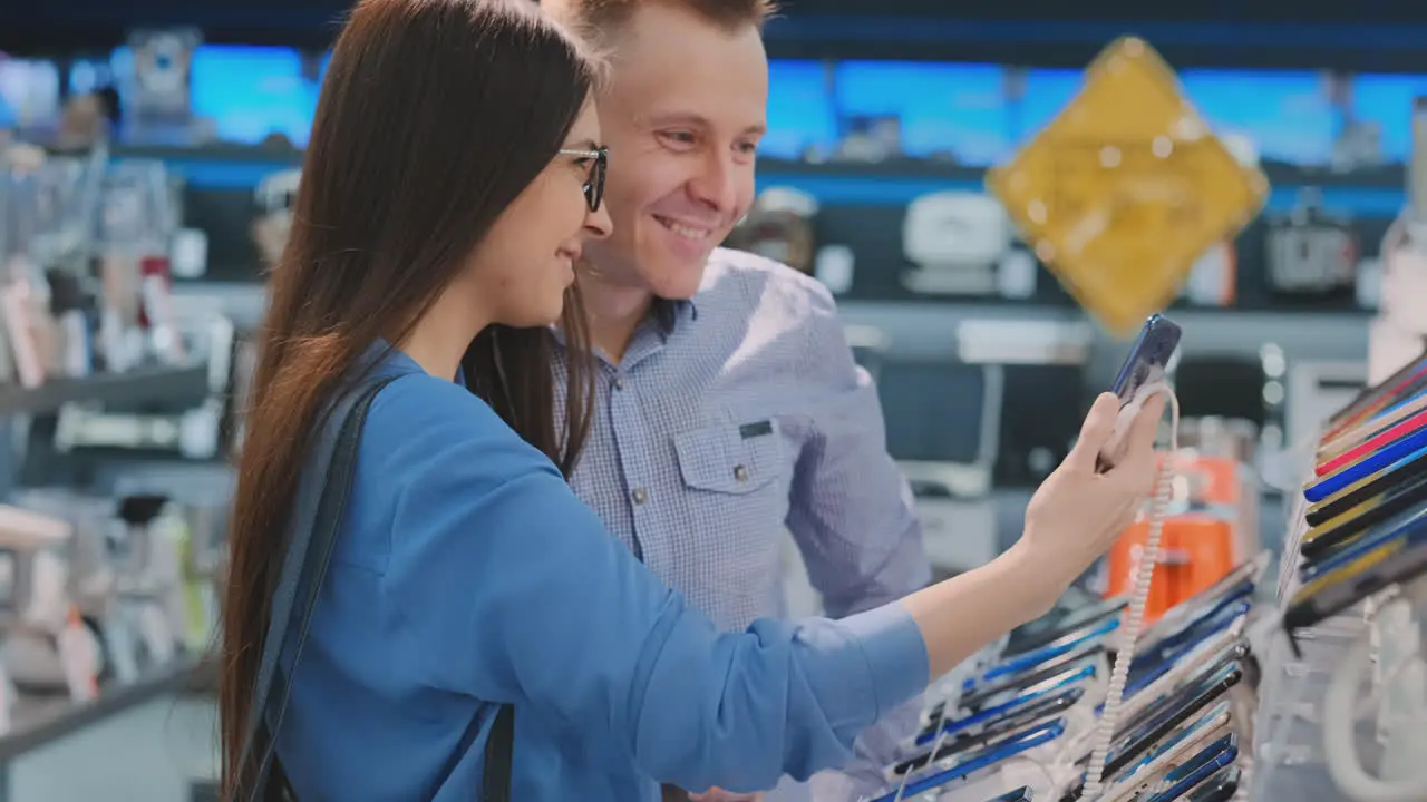 Joven Y Hermosa Pareja Casada Un Hombre Y Una Mujer Sostienen Un Teléfono Inteligente Cerca Del Escaparate Con Teléfonos Inteligentes En La Tienda De Electrodomésticos Eligiendo El Mejor Teléfono Inteligente