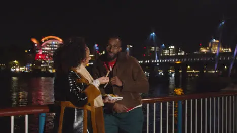 Couple On Date Standing On London South Bank At Night Eating Chips 1