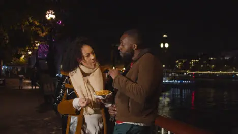 Couple On Date Standing On London South Bank At Night Eating Chips