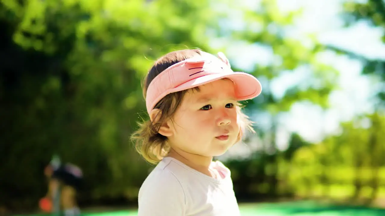 Portrait of pretty toddler girl face looking aside at the park against blurred green trees on sunny day