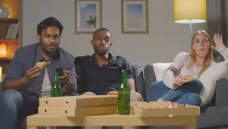 Multi-Cultural Friends Sitting On Sofa At Home Watching TV Eating Takeaway Pizza Delivery 4