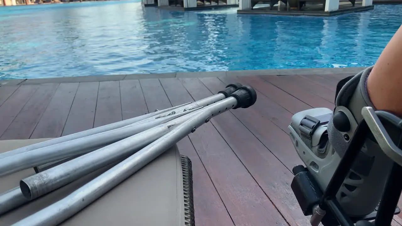 Person with a broken leg in a wheelchair and with crutches sits at the edge of a swimming pool