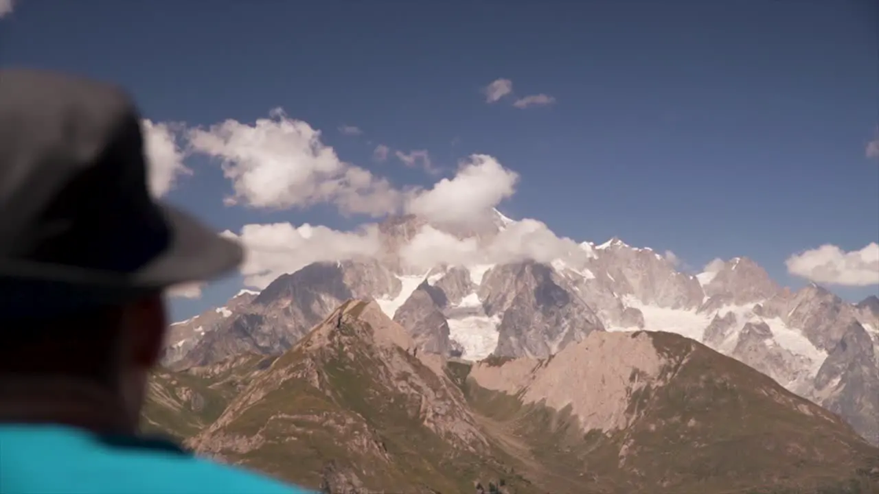 Man with a hat looking at a big mountain peak covered in clouds on a sunny day in the alps