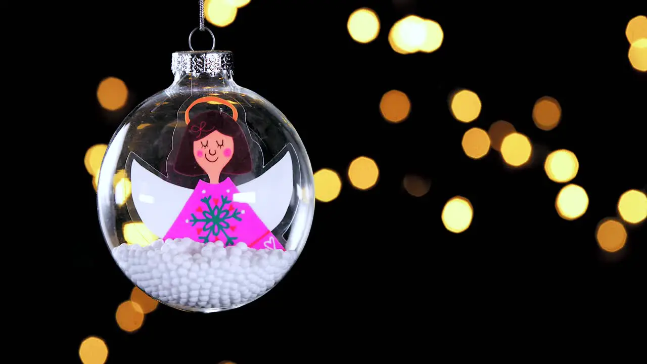 single clear Christmas ball ornament with cartoon dark hair angel and snow inside out of focus lights flickering in the background close up