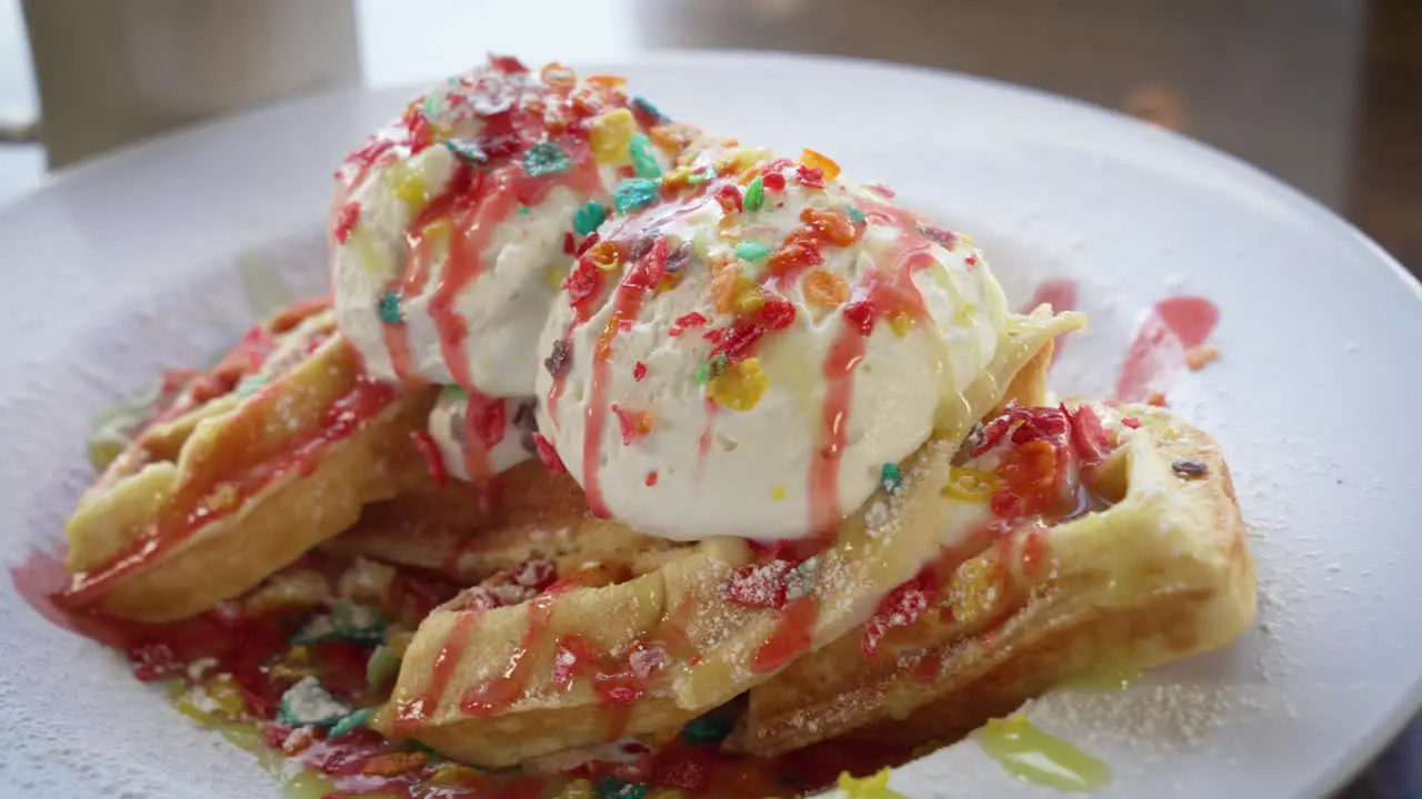 Confetti Belgian waffles filled and topped with fruity pebbles cereal and ice cream drizzled with strawberry and maple syrup close up slider 4K