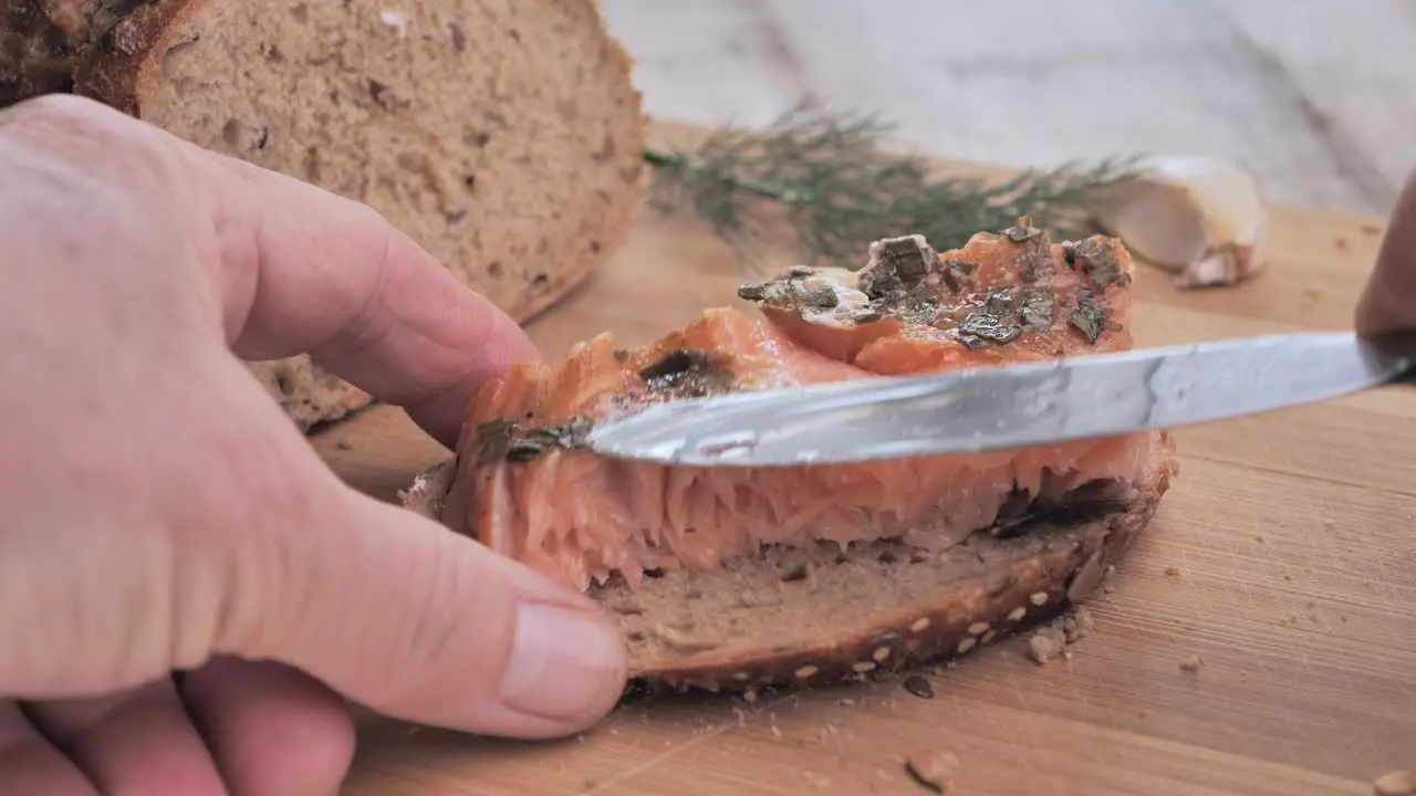 A man puts smoked salmon on a piece of bread and adds a sprig of dill on a wooden cutting board