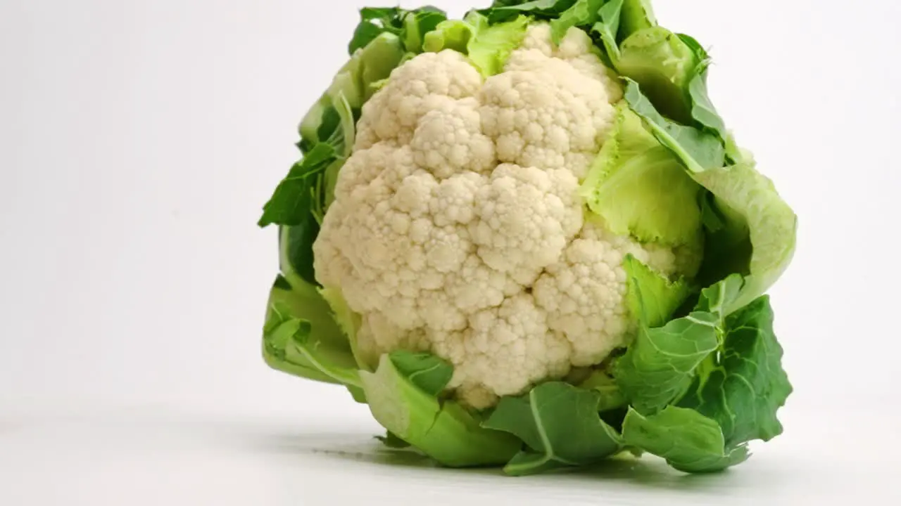 Whole head of fresh cauliflower encased in green leaves landing and bouncing on white table top in slow motion