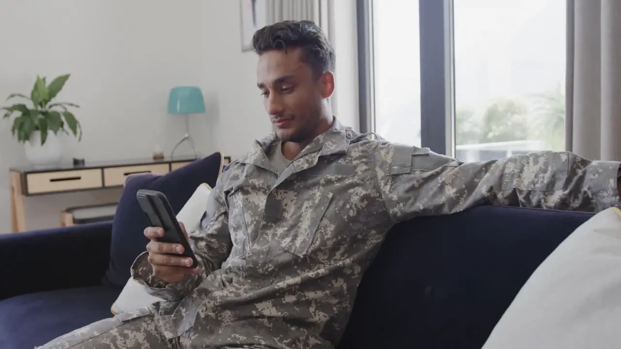 Smiling biracial male soldier in uniform sitting on sofa at home using smartphone slow motion