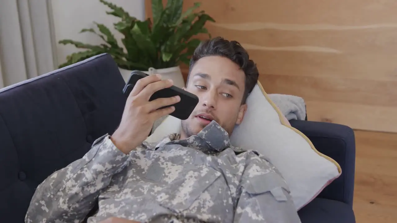 Happy biracial male soldier in uniform lying on sofa at home talking on smartphone slow motion