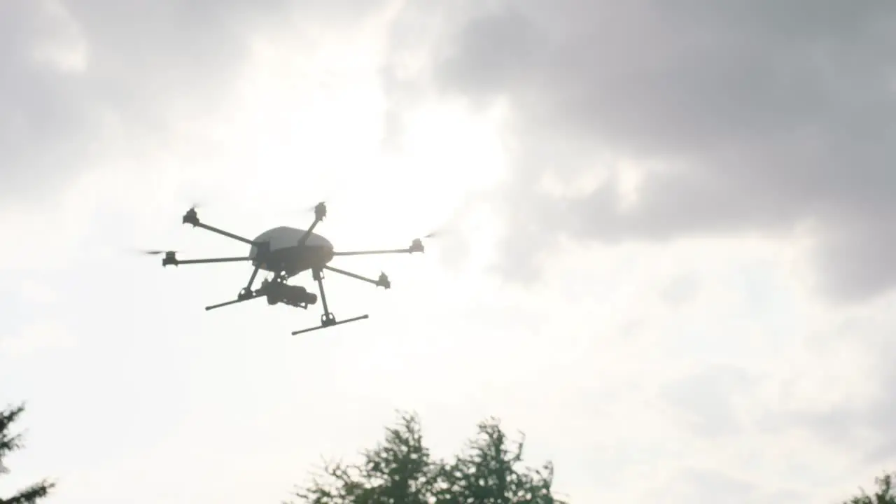 Handheld camera tracks a white drone with six propellers flying towards the right in a cloudy sky with the sun peeping through