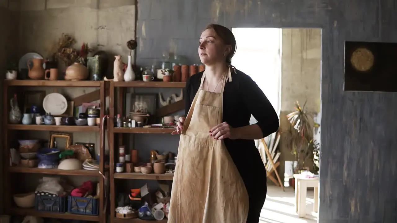 Slow Motion Footage Of Young Woman In Casual Clothes In Pottery Workshop With Ceramic Wares And Supplies On Shelves And Table With Tools Putting On Beige Apron And Smiling