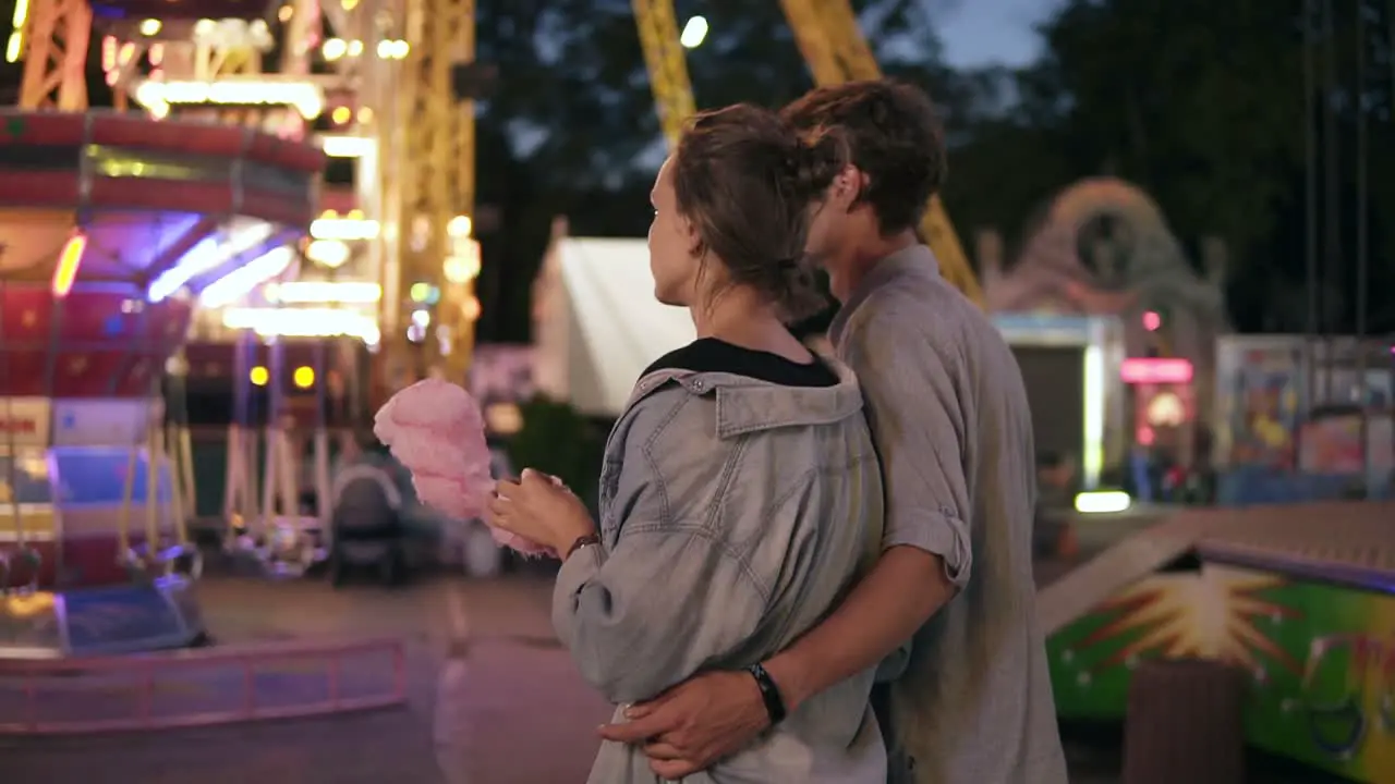 Backside View Of A Young Dating Couple Walking By Funfair At Night Eating Cotton Candy