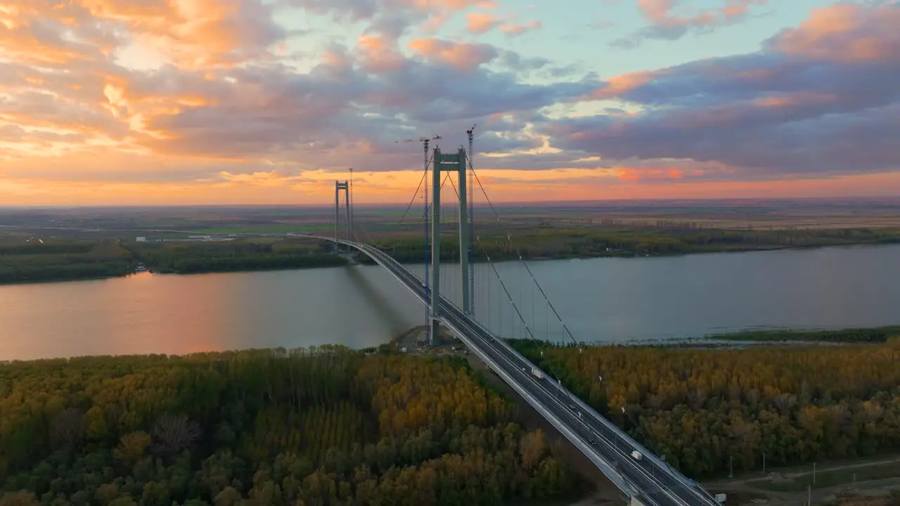 Aerial close up shot showcasing a suspended bridge spanning a vast river cars driving after sunset orange colored sky 4k50fps