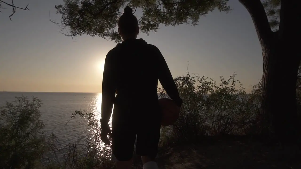 A Silhouette Of A Basketball Player Girl Comes With A Ball In Her Hand Coming Up To The Slope With Trees Around