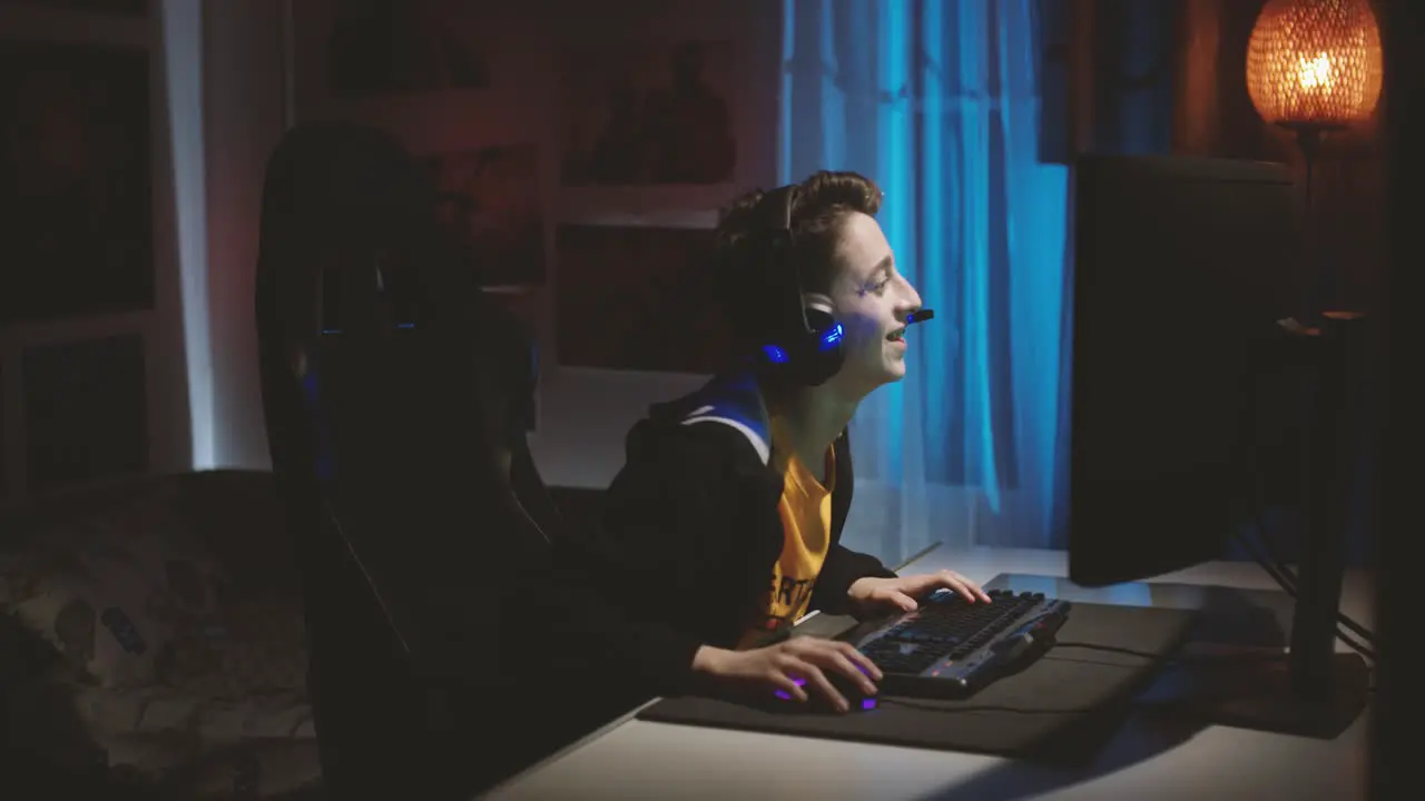 Young Caucasian Guy With Headset Talking While Playing Online Game On The Computer Inside The Room medium shot side view