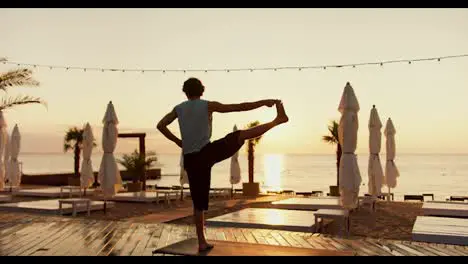 Rear view of a young guy who raised his leg up and stands on one leg on a sunny beach in the morning Yoga and stretching classes at sunrise