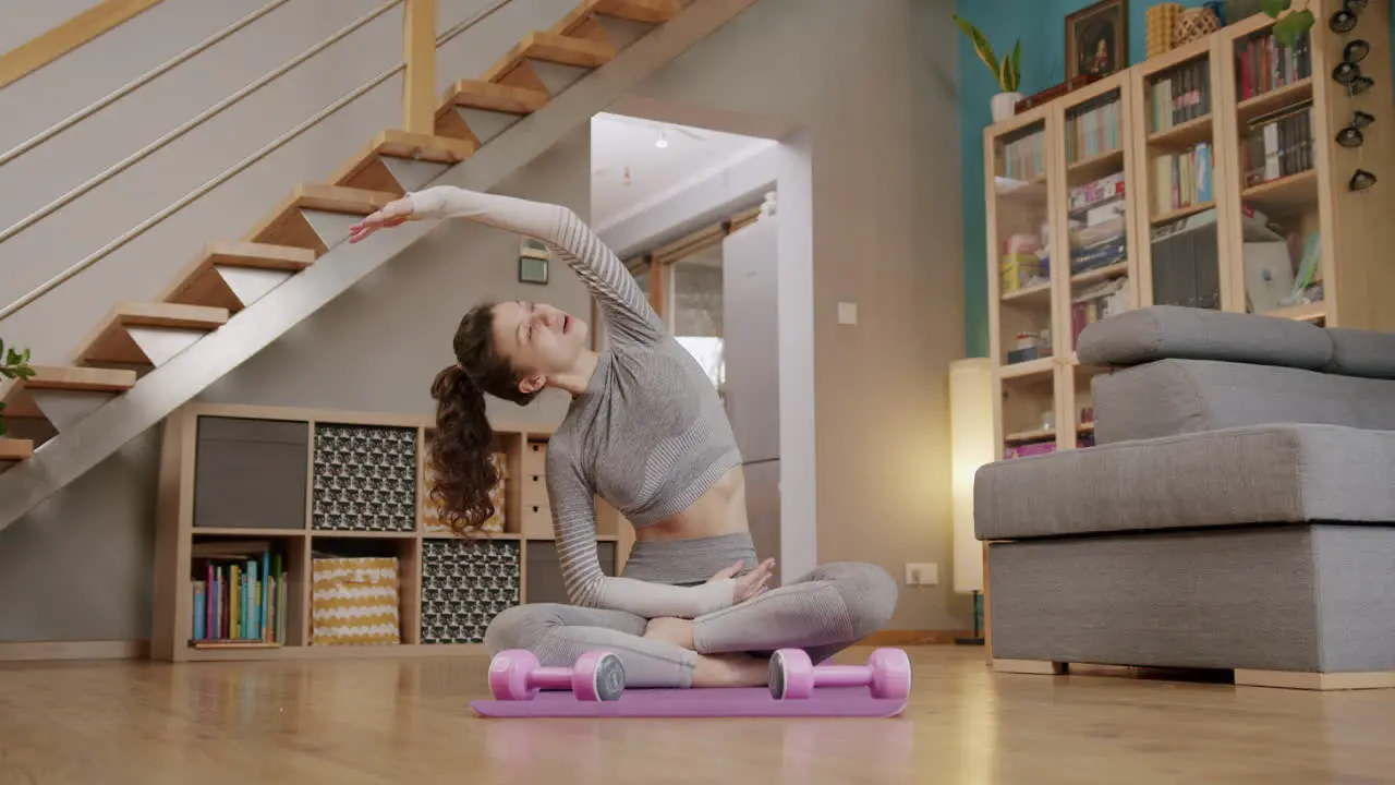 Young fit woman sits cross legged on yoga mat at home stretching with arms out