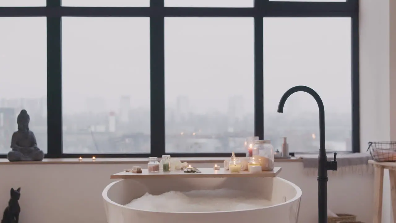 Camera Focuses On A White Bathtub With Water And Foam A Wooden Table With Bathroom Elements And A Large Window In The Background