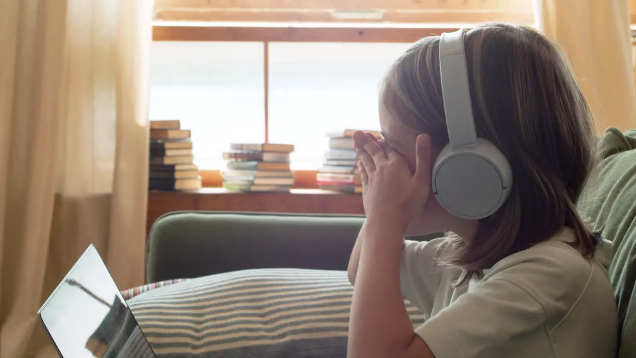 Close Up Of A Tired Little Girl With Headphones Looking At The Laptop Screen And Scratching Eyes Out Of Sleep