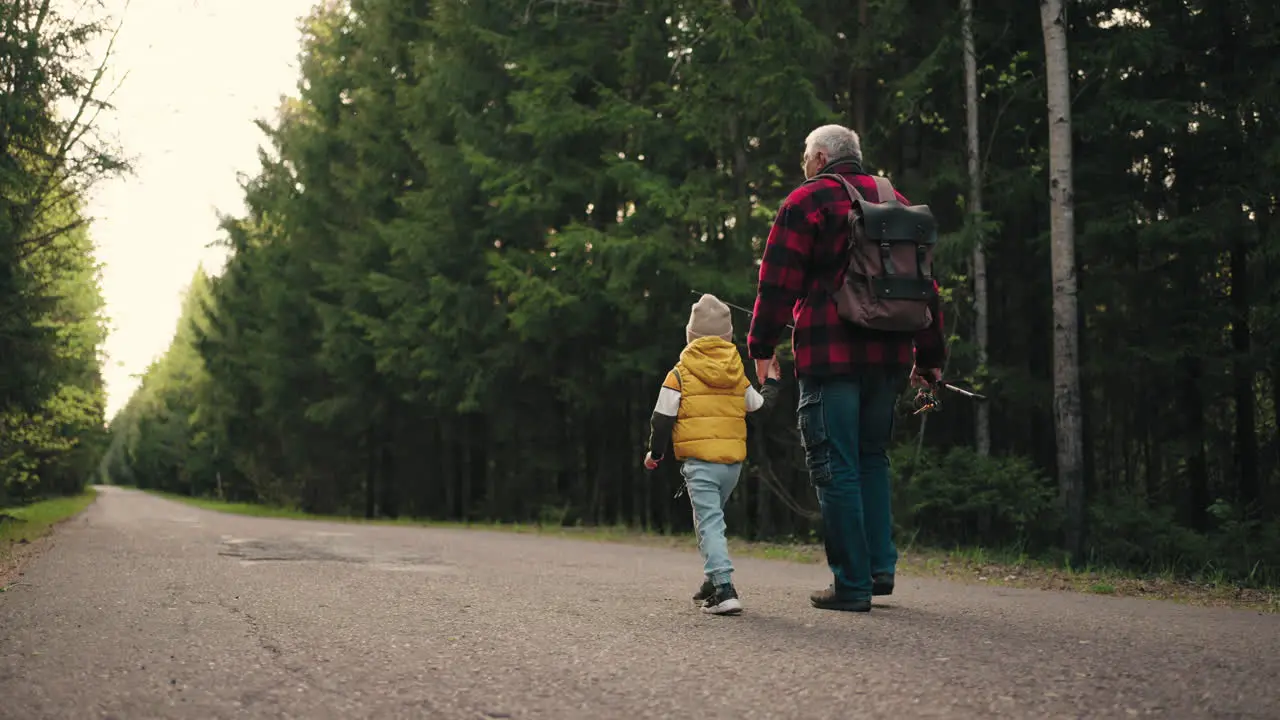 old fisherman and his little grandson are walking on road across forest in morning granddad is carrying rod