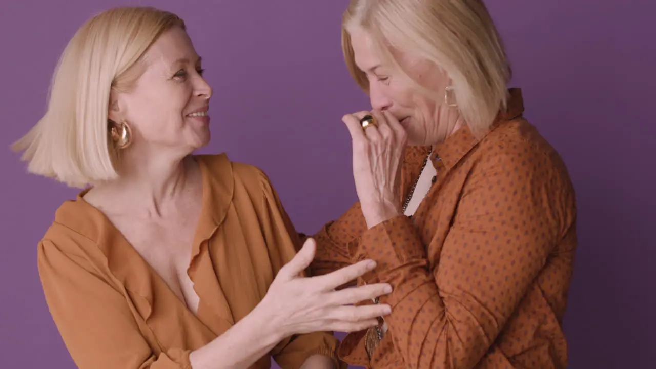 Dos Mujeres Rubias Maduras Con Pantalones Marrón Abrazándose Y Sonriendo Con Fondo Morado 1