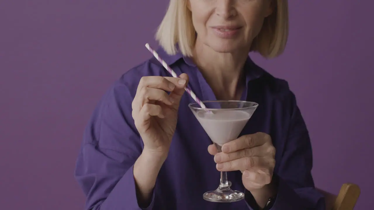 Mujer Madura Rubia Con Camisa Morada Posando Sosteniendo Un Cóctel Y Sonriendo A La Cámara Sobre Fondo Morado 1