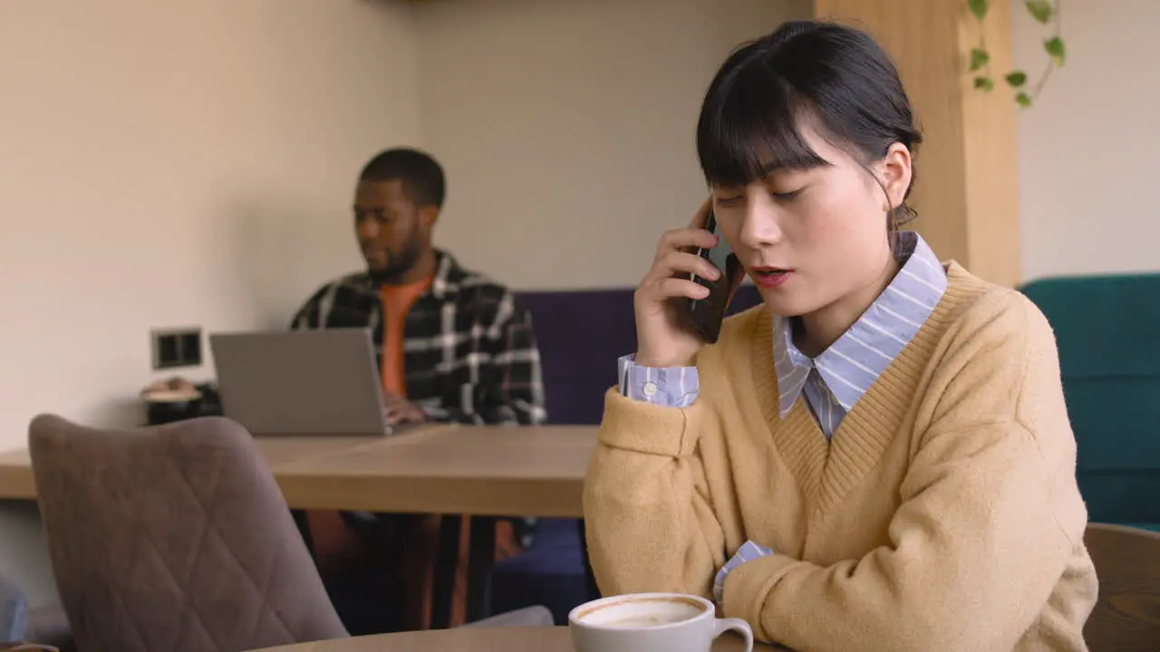 Mujer Hablando Por Teléfono Celular Sentada En Una Mesa En Una Cafetería Mientras Un Hombre Trabaja En Una Computadora Portátil Detrás De Ella 1