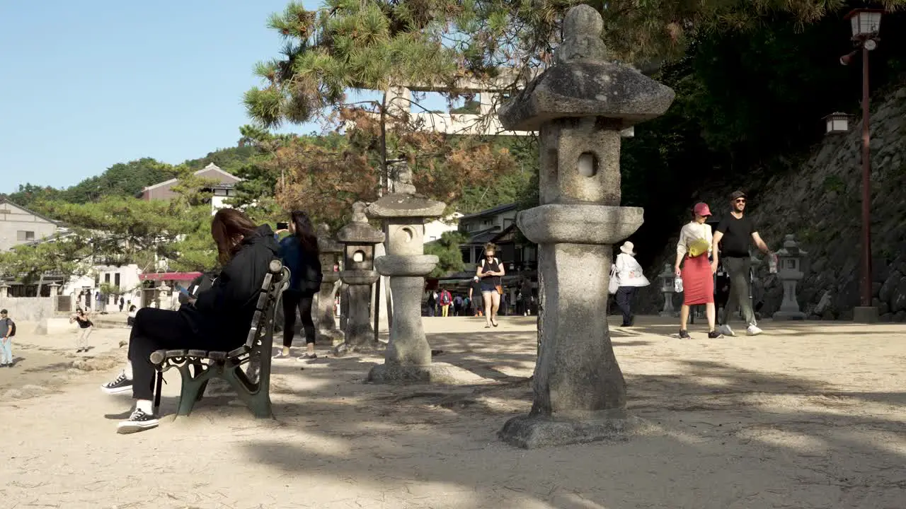 Tourist Sitting On Bench In Front Of Row Of Stone Lanterns At Itsukushima Shrine On Sunny Afternoon