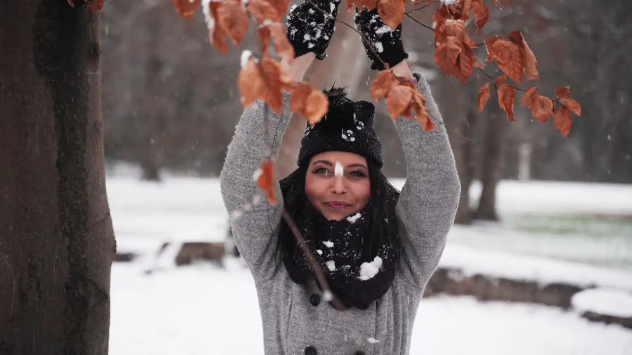 woman in the winter in a park holding hands up and dropping snow on her while smiling with leaves in front mid shot