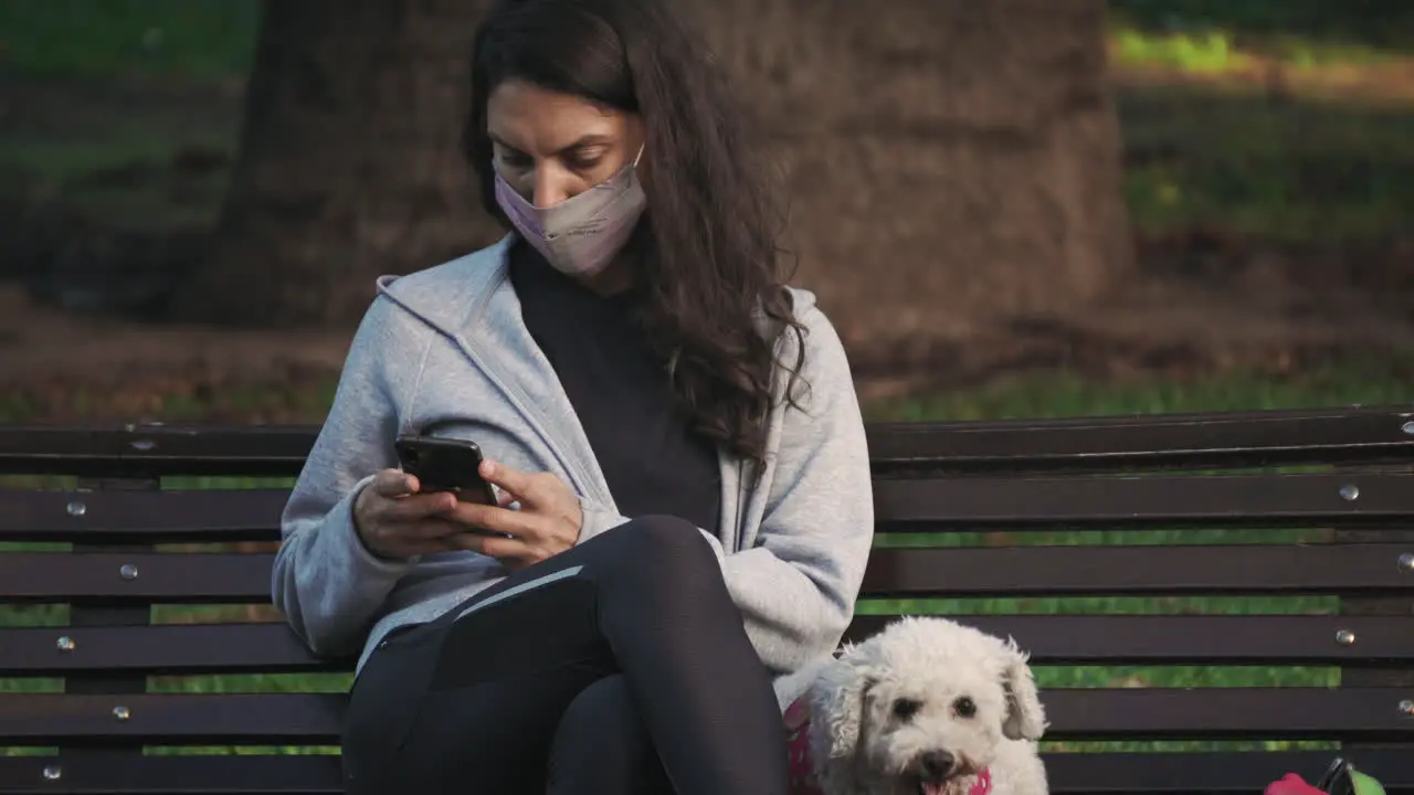 Woman wearing a mask sitting by her dog on park bench using her phone