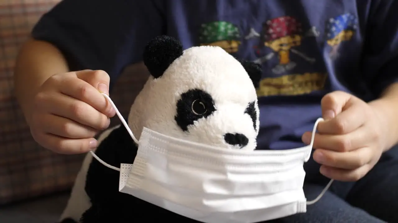 Young Caucasian boy puts mask on stuffed baby panda Covil-19 quarantine