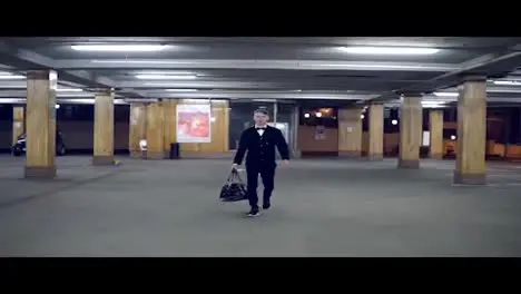 Young blonde man in a black suit with bow-tie walking on the parking with big leather bag Slow Motion