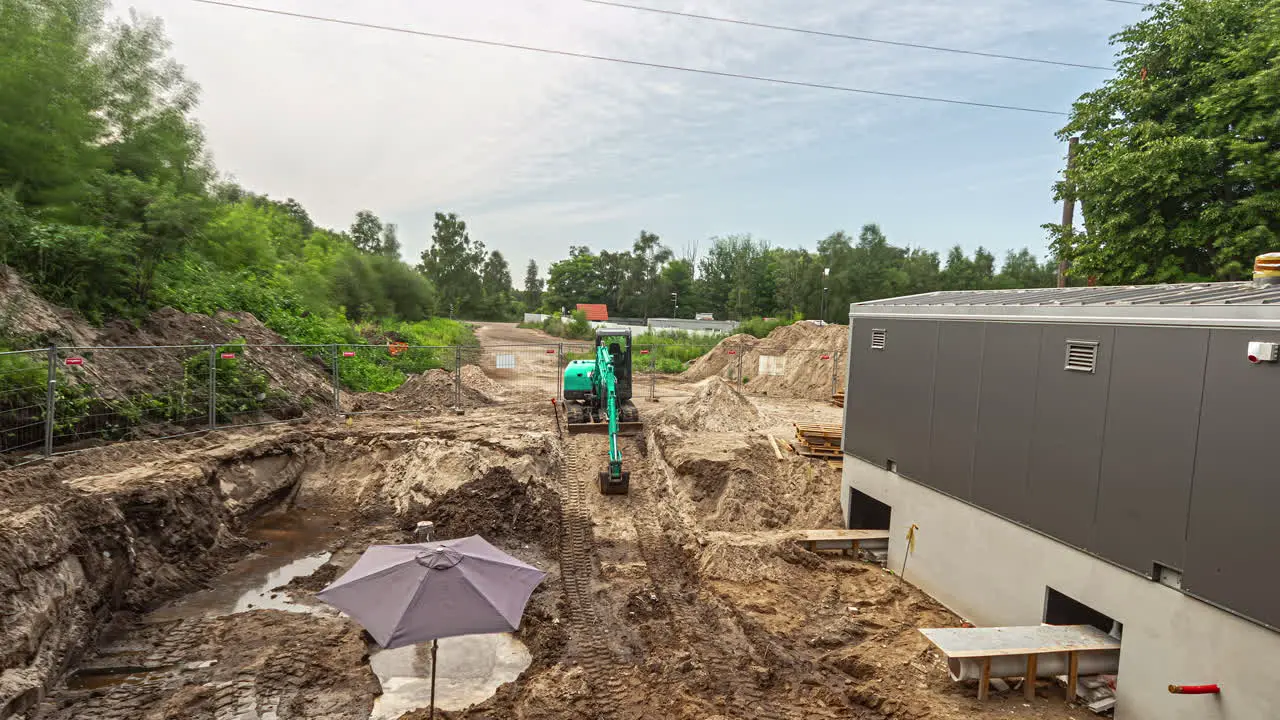  High angle shot of green excavator leveling earth for construction of a parking lot beside newly made building throughout the day in timelapse