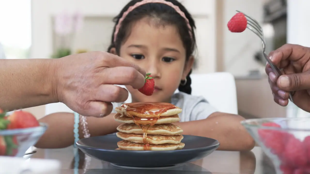 Biracial girl enjoys breakfast as hands help her with syrup and fruit