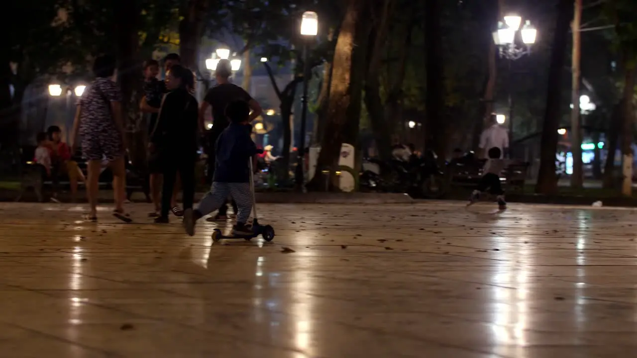 Kid rides glider scooter while his family watches from a nearby bench in a local park at night Low angle tracking handheld shot