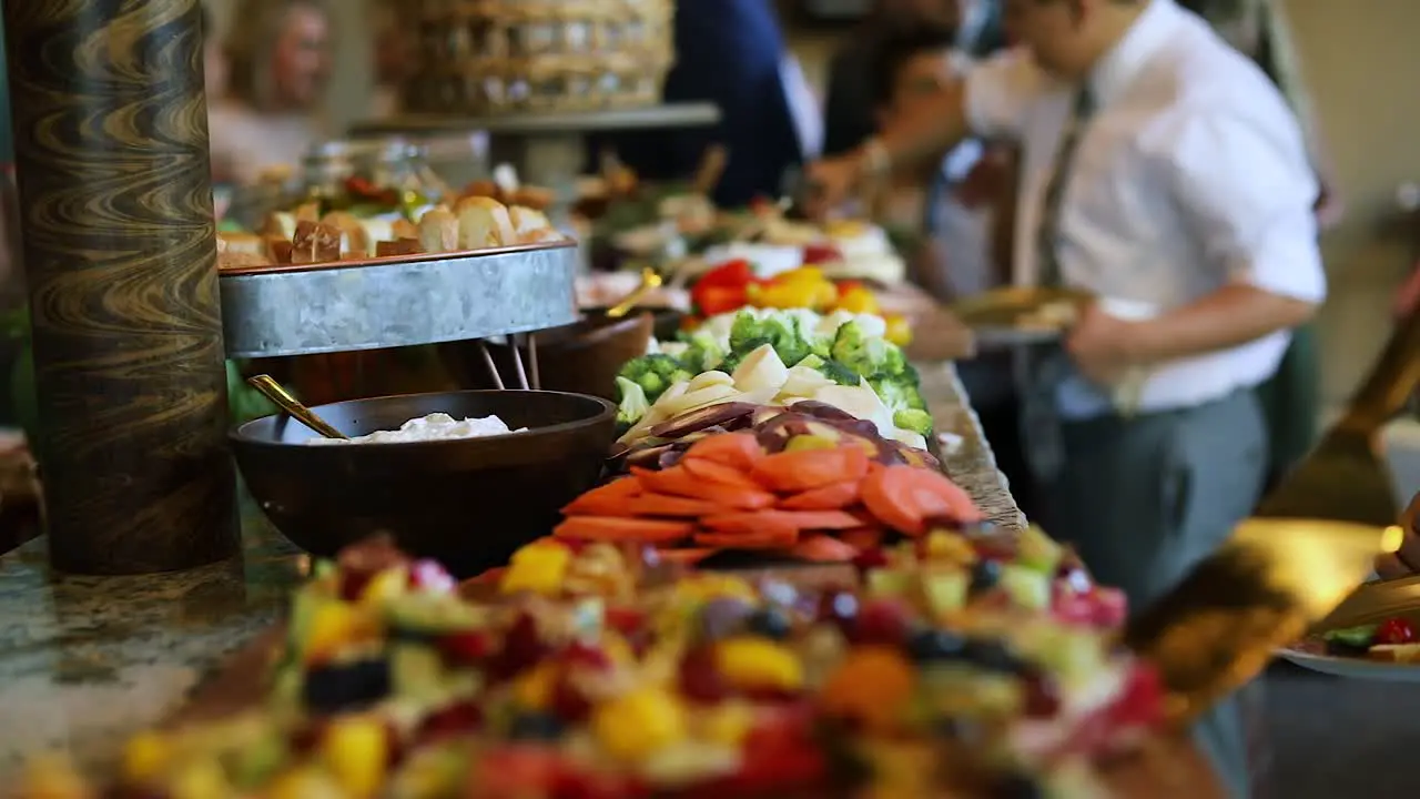 Smooth shot of a line of people at a crowded event going through a food line and grabbing a variety of food including meats cheeses bread fruits and vegetables